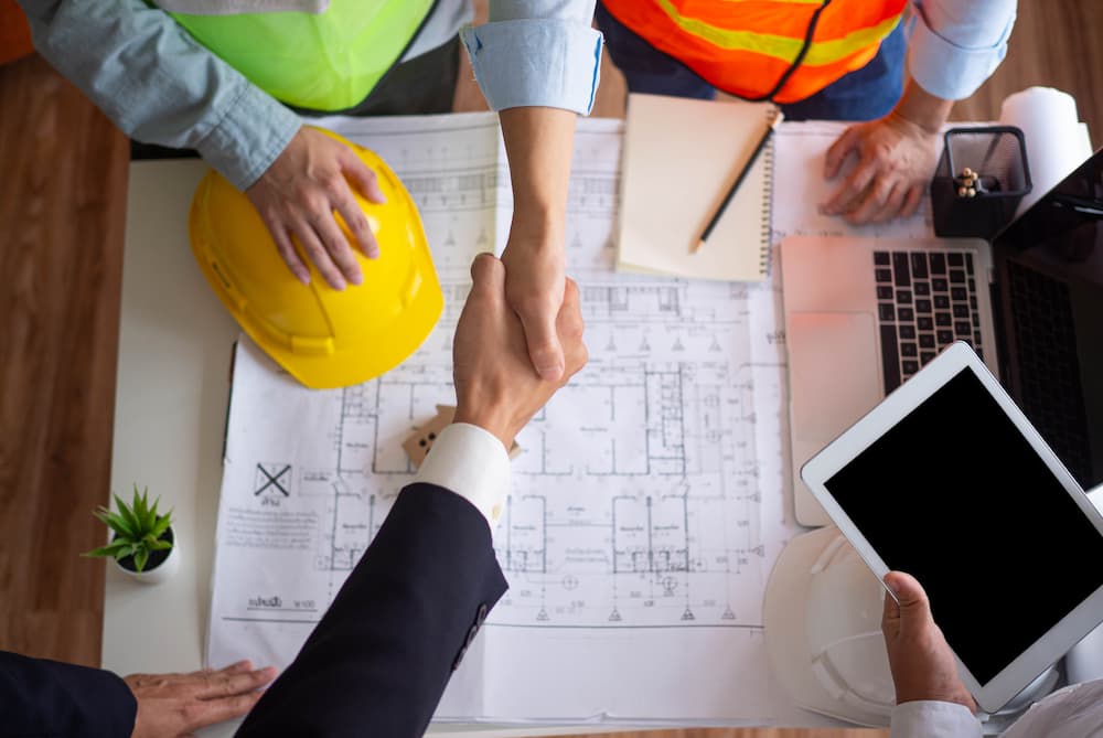 Overhead view of a contractor and client shaking hands over architectural blueprints, providing tips on how to choose a contractor for home renovations.